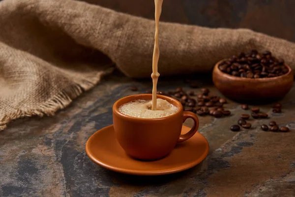 Наливая горячий кофе в чашку на блюдце на мраморной поверхности возле мешковины и бобов — стоковое фото