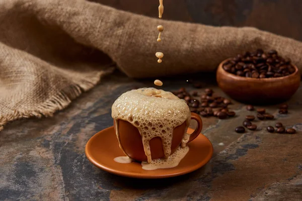 Derramando café quente em xícara com espuma na superfície de mármore perto de pano de saco e feijão — Fotografia de Stock