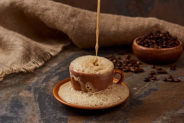 Versando caffè caldo in tazza con schiuma sul piattino sulla superficie di marmo vicino a sacco e fagioli — Foto stock