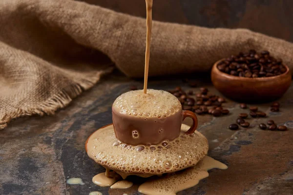 Fluxo de café quente na xícara com espuma e poça no pires na superfície de mármore perto de pano de saco e feijão — Fotografia de Stock