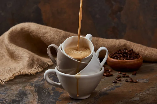Fluxo de café quente a partir de copos na superfície de mármore perto de pano de saco e feijão — Fotografia de Stock