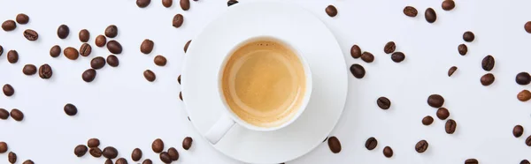 Панорамний знімок смачної кави в чашці біля розкиданих обсмажених бобів на білому тлі — стокове фото