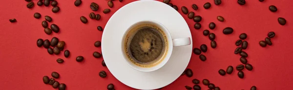 Panoramaaufnahme von köstlichem Kaffee in Tasse in der Nähe verstreuter gerösteter Bohnen auf rotem Hintergrund — Stockfoto