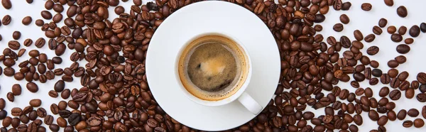 Панорамный снимок вкусного кофе в коричневой чашке возле разбросанных жареных бобов на белом фоне — стоковое фото