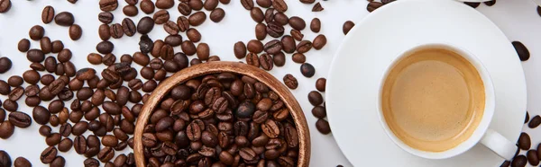 Верхний вид кофе в чашке на блюдце рядом с разбросанными жареными бобами и деревянной миской, панорамный снимок — стоковое фото