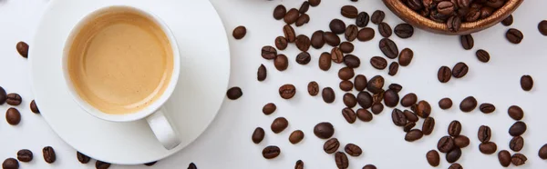Вид сверху на кофе в чашке на блюдце рядом с разбросанными жареными бобами, панорамный снимок — стоковое фото