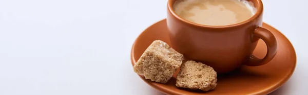 Plan panoramique de café savoureux avec mousse dans une tasse sur soucoupe près de sucre brun sur fond blanc avec espace de copie — Photo de stock