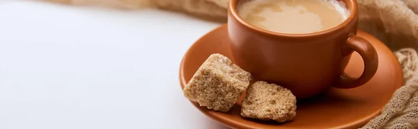 Panoramaaufnahme von leckerem Kaffee mit Schaum in Tasse auf Untertasse in der Nähe von braunem Zucker auf weißem Hintergrund mit Tuch — Stockfoto