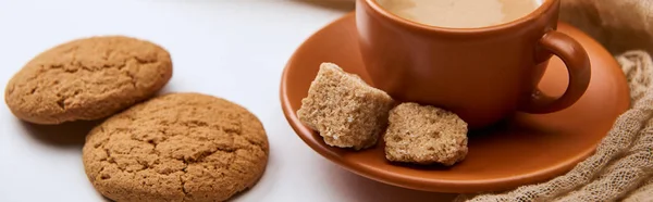 Panoramic shot of delicious coffee with foam in cup on saucer near brown sugar and cookies on white background — Stock Photo