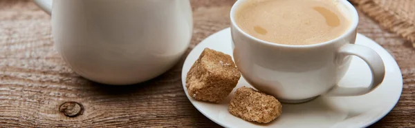 Tiro panorâmico de café delicioso com espuma na xícara no pires com açúcar mascavo perto de pano de saco e jarro de leite no fundo de madeira — Fotografia de Stock