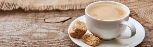 Panoramaaufnahme von köstlichem Kaffee mit Schaum in Tasse auf Untertasse mit braunem Zucker in der Nähe von Sacktuch auf Holzgrund — Stockfoto