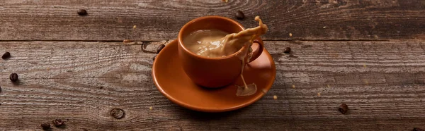Tiro panorâmico de respingo de café em xícara marrom na mesa de madeira com grãos de café espalhados — Fotografia de Stock