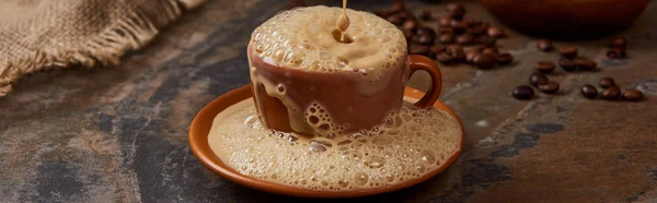 Panoramic shot of coffee foam with bubbles pouring out of cup on saucer on marble surface — Stock Photo