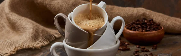 Панорамный снимок вытекающего горячего кофе из чашек возле мешковины — стоковое фото