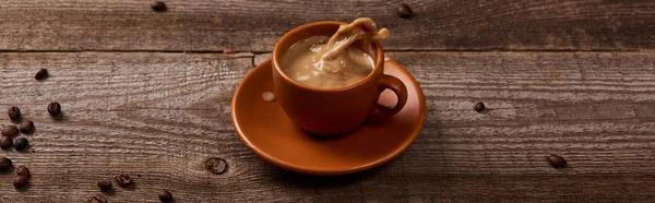 Plan panoramique d'éclaboussure de café dans une tasse sur une table en bois avec des grains de café éparpillés — Photo de stock