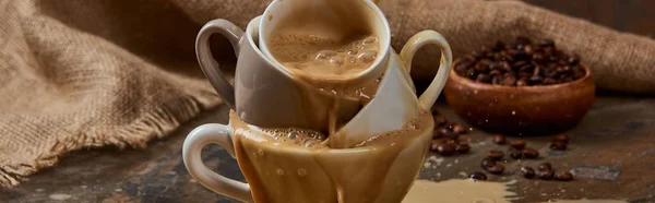Панорамный снимок вытекающего горячего кофе из чашек на мраморном столе возле мешковины и бобов — стоковое фото