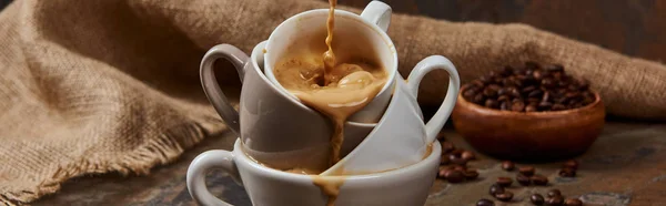 Панорамный снимок вытекающего горячего кофе из чашек на мраморной поверхности возле мешковины и бобов — стоковое фото