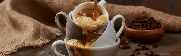 Панорамный снимок вытекающего горячего кофе из чашек на мраморном столе возле мешковины и бобов — стоковое фото