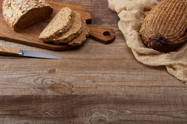Frisch gebackenes geschnittenes Brot mit Samen auf Schneidebrett neben Messer und Brot auf Tuch auf Holztisch — Stockfoto