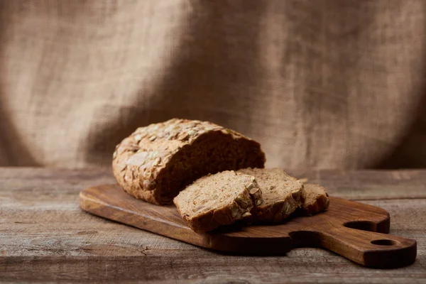 Вкусный нарезанный коричневый хлеб на деревянной доске с мешковиной на заднем плане — стоковое фото