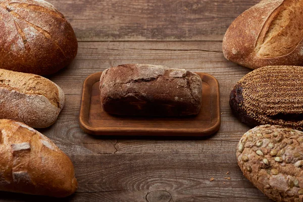 Delicious loaves of bread around brown bread on board on wooden table — Stock Photo