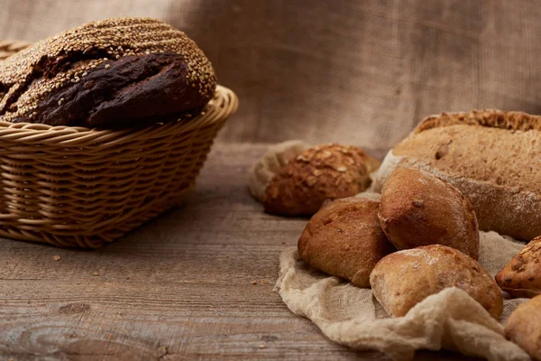 Вкусные свежие булочки и хлеб в плетеной коробке на деревянном деревенском столе с тканью — стоковое фото