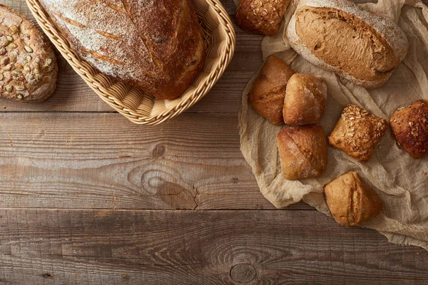 Top view of fresh baked bread in wicker basket and buns on cloth on wooden table — Stock Photo