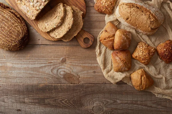 Вид сверху на свежие ломтики хлеба на доске и булочки на ткани на деревянном столе — стоковое фото