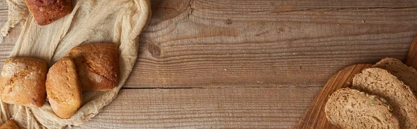 Вид сверху на свежие ломтики хлеба на доске и булочки на ткани на деревянном столе, панорамный снимок — стоковое фото