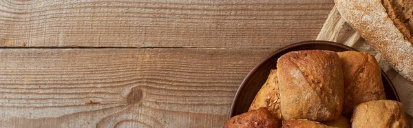Vista superior de pan fresco y bollos en tazón sobre tela sobre mesa de madera, plano panorámico - foto de stock