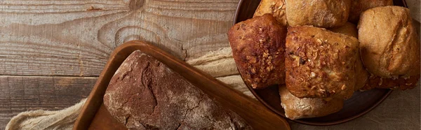 Draufsicht auf frische Brotlaibe auf Schneidebrett und Brötchen in Schüssel auf Tuch auf Holztisch, Panoramaaufnahme — Stockfoto