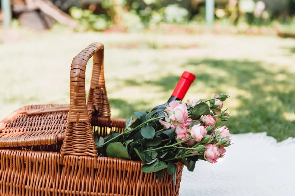 Weidenkorb mit Rosen und einer Flasche Wein auf einer weißen Decke an einem sonnigen Tag im Garten — Stockfoto