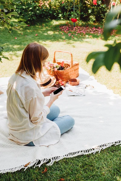 Блондинка сидить на ковдрі в саду і має пікнік в сонячний день, використовуючи смартфон — стокове фото