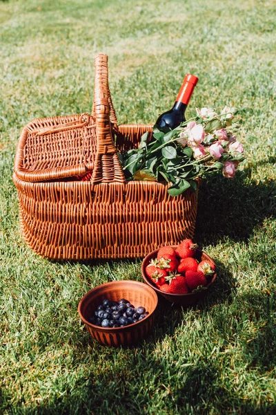 Wicker basket with roses and bottle of wine on green grass near strawberries and blueberries in bowls — Stock Photo