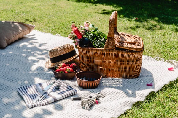 Cesta de vime com rosas e vinho em cobertor branco perto de chapéu de palha, talheres em guardanapo e bagas no dia ensolarado no jardim — Fotografia de Stock