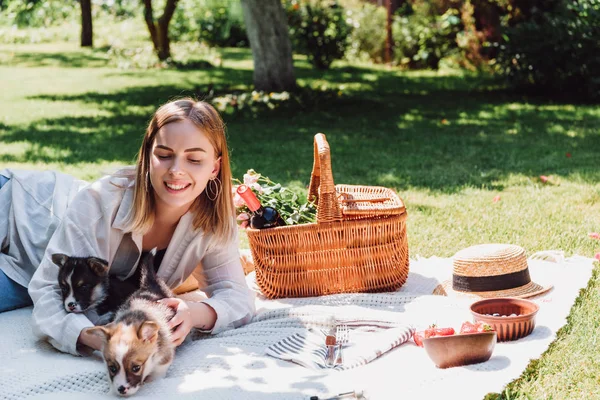 Улыбающаяся блондинка сидит на одеяле в саду и устраивает пикник с щенками в солнечный день — стоковое фото