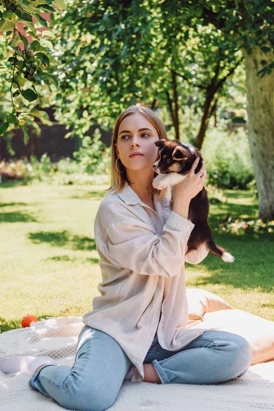 Fille blonde avec les yeux fermés tenant chiot mignon dans le jardin vert et regardant loin — Photo de stock