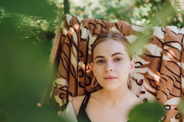 Selective focus of green leaves and beautiful blonde girl sitting in deck chair in garden — Stock Photo