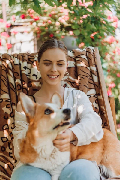 Enfoque selectivo de la chica rubia feliz sosteniendo perro corgi mientras está sentado en la silla de cubierta en el jardín - foto de stock