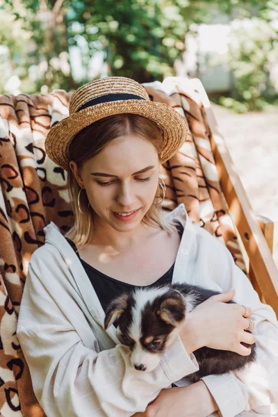 Sonrisa rubia chica en paja sombrero celebración cachorro mientras sentado en cubierta silla en jardín - foto de stock