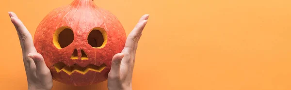 Cropped view of woman holding carved spooky Halloween pumpkin on orange background, panoramic shot — Stock Photo