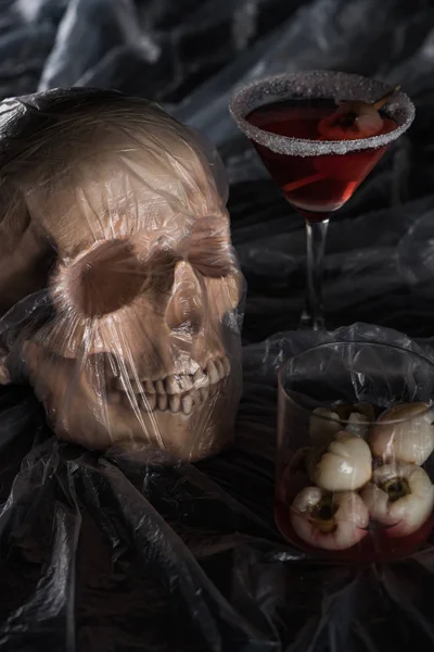 Spooky human skull in cellophane near red cocktail on black background, Halloween decoration — Stock Photo