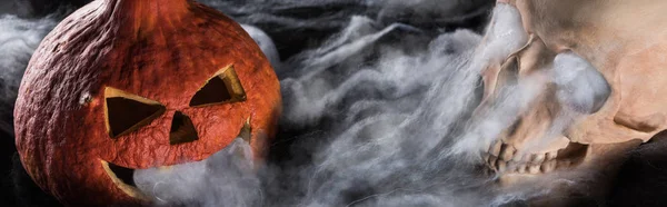 Panoramic shot of spooky human skull and carved Halloween pumpkin on black background — Stock Photo