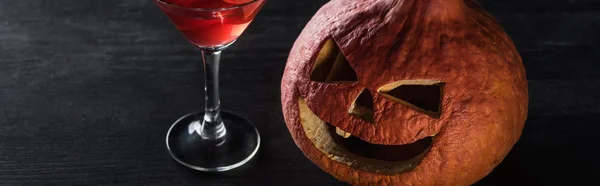 Panoramic shot of carved Halloween pumpkin and red cocktail on black background — Stock Photo