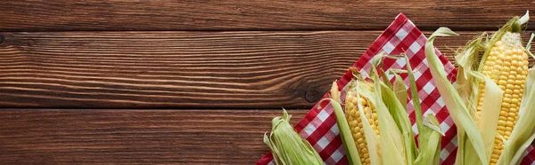 Panoramic shot of raw corn on checkered tablecloth on wooden surface — Stock Photo