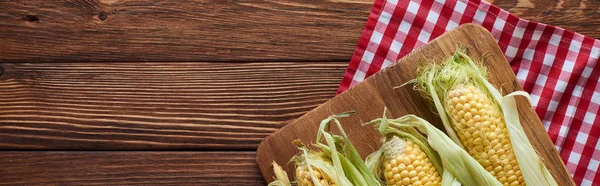 Panoramic shot of cutting board with raw corn on checkered tablecloth on wooden surface — Stock Photo