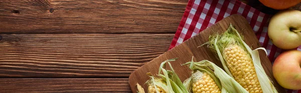 Panoramic shot of cutting board with raw corn near apples on checkered tablecloth on wooden surface — Stock Photo