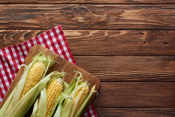 Top view of cutting board with raw corn on checkered tablecloth on wooden surface — Stock Photo