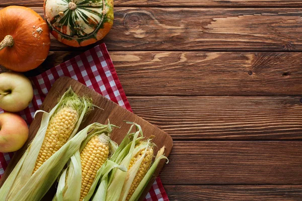 Top view of checkered tablecloth with apples, pumpkins and cutting board with corn on wooden surface with copy space — Stock Photo