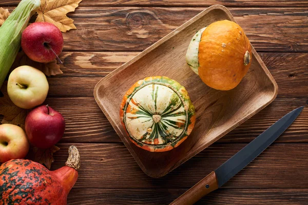 Vista dall'alto di piatto rettangolare marrone con zucche fresche vicino a coltello e mele su superficie di legno con foglie secche — Foto stock